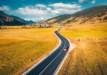 aerial view of the road in mountain valley at sunset in autumn. top view of asphalt roadway, car, hills with orange grass, blue sky, trees, buildings. highway and fields in fall. colorful landscape