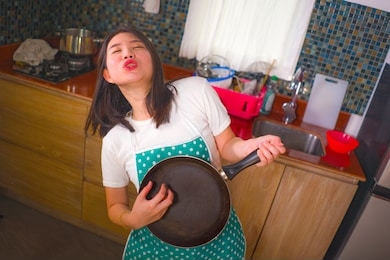 natural lifestyle portrait of young beautiful and happy asian korean woman in kitchen apron playing air guitar with cooking pan having fun enjoying domestic chores dancing and singing at home