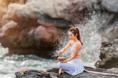 beautiful asian woman is practicing yoga meditation by tibetan singing bowl on the asana in outdoors with sea and wave on background. healthcare and exercise concept.