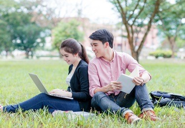female and male students sitting on the green field and looking at laptop computer with happily 