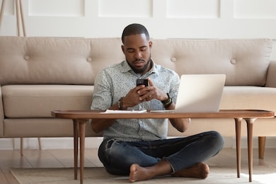 african guy sits leaned on couch cross-legged on carpet near coffee table hold cellphone distracted from work chat online, self-employed man solve business issues contact with client distantly concept