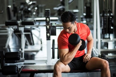 strong asian muscle man sit and exercise biceps with heavy dumbbells with gym equipment background. muscular body sportsman in sport fitness center. bodybuilding and healthy lifestyle concept.