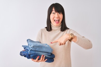 beautiful chinese shopkeeper woman holding folded jeans over isolated white background very happy pointing with hand and finger