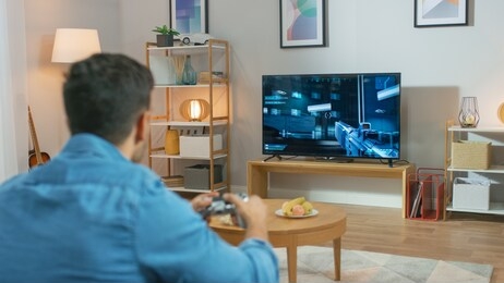 in the living room man sitting on a couch holds controller playing in a console video game, 3d action shooter gameplay shown on tv screen.