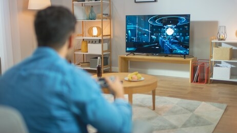 in the living room man sitting on a couch holds controller playing in a console video game, 3d action shooter gameplay shown on tv screen.