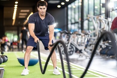 sporty asian man exercising with battle ropes at the gym on green floor. strong male determine with her indoor workout for stamina and building muscular body. athlete battle rope workout concept.