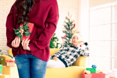 a beautiful asian woman is surprising his boyfriend by hiding a gift box in the back and walking towards him while the man is lying on the sofa in the room with a christmas tree behind them.
