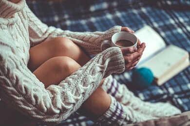 comforting cozy woman in knitted winter socks and white warm sweater sitting on a plaid blanket and warming up with a cup of hot cocoa at home in winter time. cozy time and winter drinks 
