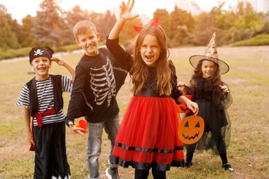 girl in devil costume holding paper jack o lantern and scaring camera while having fun with friends during halloween masquerade in park