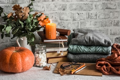 autumn cozy home still life. pumpkin, stack of books, burning candle, pile of winter autumn sweaters, jar of marshmallows on the kitchen table. flat lay