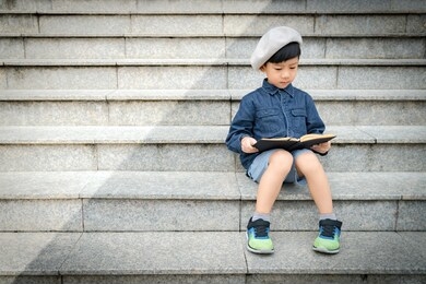 portrait of a cute little asian schoolboy  in stylish outfit with beret sitting on a stairs in front of a library concentration reading a story book. child’s brain development, cognitive skills.