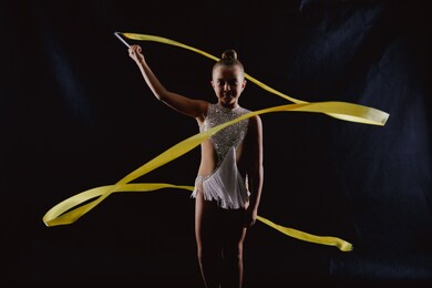 girl gymnast in a white sparkling gymnastic swimsuit makes a snake in the air with a yellow ribbon on a black background