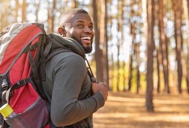 smiling young african man wearing backpack standing on autumn forest trail, looking aside, hiking alone, copy space