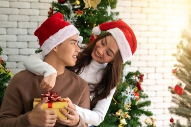 young asian adult teenager couple holding gift present box hugging and celebrateing christmas holiday together in living room with christmas tree ornament decoration.