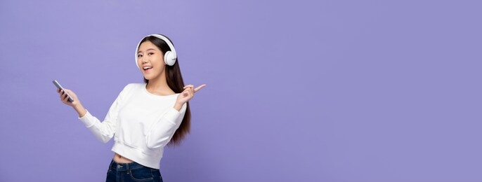 young pretty asian woman wearing headphones listening to music from mobile phone and pointing to copy space on light purple banner background