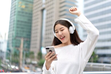 young happy asian girl wearing headphones listening to music online from smartphone and raising hand up in city building background