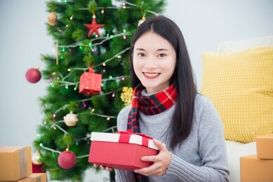 beautiful young asian girl sitting on floor at living room with christmas tree decoration and holding gift box and smiles at camera.
