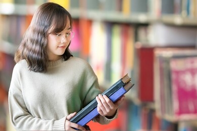 happy asian woman looking for a book in a library. woman reading book in an university library. library and education concept.