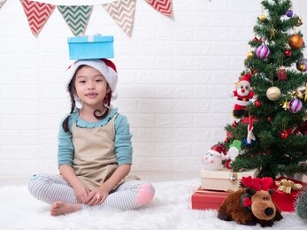 asian little cute girl 6 years old sitting on carpet and put a gift box on her head in white room with the christmas tree. christmas celebration concept.