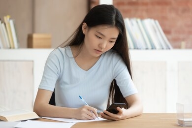 asian schoolgirl do homework seated at library desk, focused student girl hold cell phone read info noting get helpful information from internet prepare thesis, study use websites distant help concept
