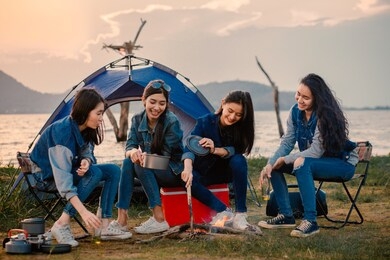a group of young women camping, they sit around the fire, cook and party together. background it is a large river and mountain camping and tarvel concept.