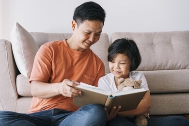 asian father and daughter reading book while sitting in the living room at her home.