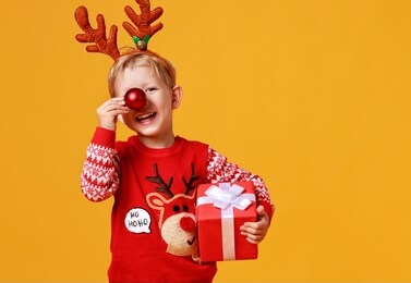 happy funny child boy in red christmas reindeer costume with gift on yellow colored background
