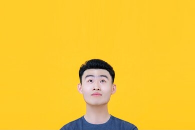 young handsome clean shaven asian man with thoughtful facial expression looking up to copy space with raised eyebrows isolated on yellow background