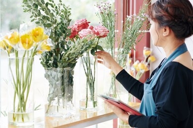 professional florist checking order on tablet computer and choosing flowers from vases on shop window sill