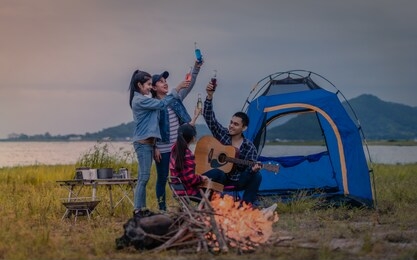 group of asian young friends camping by playing guitar and drinking beverage to happily near tent and camp fire with lake background. lifestyle and camping concept.