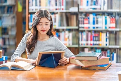 asian young student in casual suit doing homework and using technology teblet in library of university or colleage with various book and stationary over the book shelf background, back to school