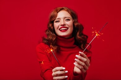  girl with sincere smile looks into camera, posing with burning christmas sparklers. portrait of redhead lady in red sweater on isolated background