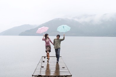 joyful and spontaneous japanese couple in love, holding hands and running on a wooden pier on a natural lake and mountains landscape, having fun and laughing outdoors.