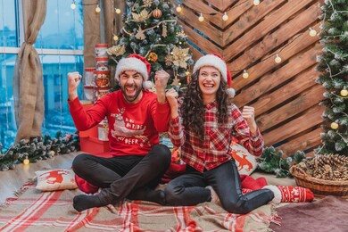 extremely happy man and woman are raising clunched fists, saying yes, while sitting near christmas tree in lotos position.