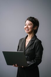studio portrait of a confident, young, professional and attractive business woman in a black dress smiling as she looks up from typing on her laptop computer against a white / silver background. 