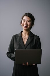 studio portrait of a confident, young, professional and attractive business woman in a black dress smiling as she looks up from typing on her laptop computer against a white / silver background. 