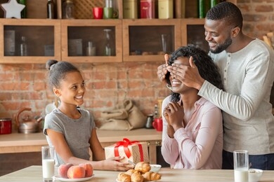 birthday, mother day concept. cheerful african little girl holding gift for mother, smiling man closing woman eyes, kitchen interior, free space