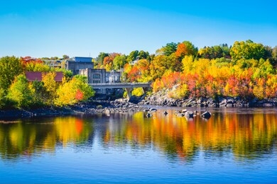autumn landscape view of color trees with dam on lake in city park in chicoutimi saguenay quebec canada. beautiful fall foliage on lakeside reflection in water. natural colorful scenery in fall season