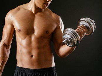 photo of an asian male exercising with dumbbells and doing bicep curls over dark background.