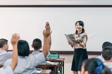 an smiling asian female high school teacher teaches the white uniform students in the classroom by asking questions and then the students raise their hands for answers.