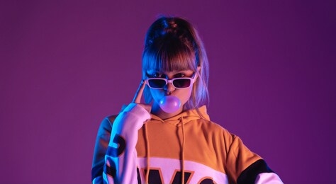 pretty young 20s fashion teen girl model wear glasses blowing bubble gum looking at camera standing at purple studio background, igen teenager in trendy stylish night glow 80s 90s concept, portrait