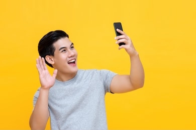 young smiling  handsome asian man saying hi and waving hand while making video call on smarphone isolated on yellow background