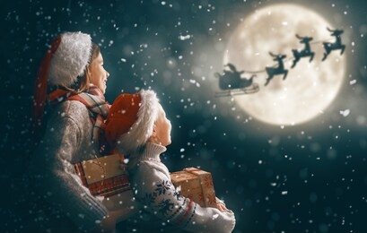 merry christmas and happy holidays! cute little children with xmas presents. santa claus flying in his sleigh against moon sky. kids enjoying the holiday with gifts on dark background.