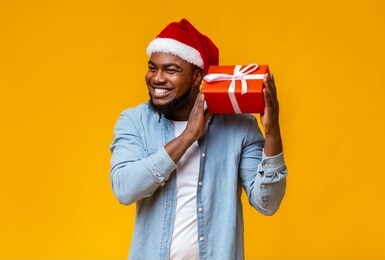 guess what's inside. happy black man in santa hat shaking christmas present, listening what rattles in gift box, yellow background