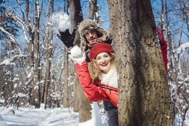 woman and man in winter throwing snowball at the camera hiding behind a tree