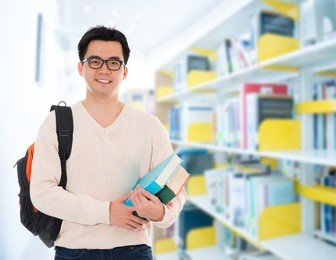 asian adult student in casual wear with school bag carrying text books standing inside school library building.
