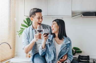 happy asian couple in love laughing and drinking red wine in the kitchen to celebrate their anniversary