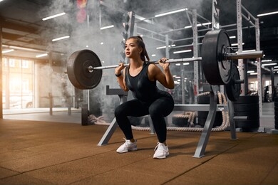female weightlifter is getting ready to stand with heavy barbell, squatting with weight, looking away with pleasant expression, enjoying training in gym, portrait, side shot