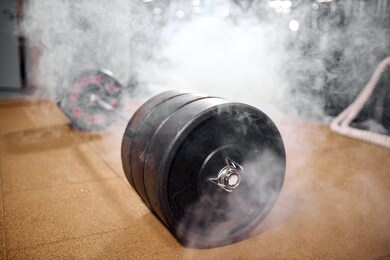 black metal heavy barbell in white smoke, standing on brown floor in modern gym hall, bodybuilder, sports people concept, close up, indoor shot