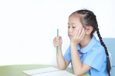 unhappy asian little schoolgirl studying and tired with doing her homework on desk over white background. kid girl hold and looking pencil in her hand with thinking something.